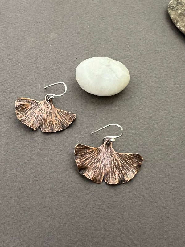 Copper leaf-shaped earrings on a gray surface with a white stone. Organic style and earrings, medium brown with silver ear wires.