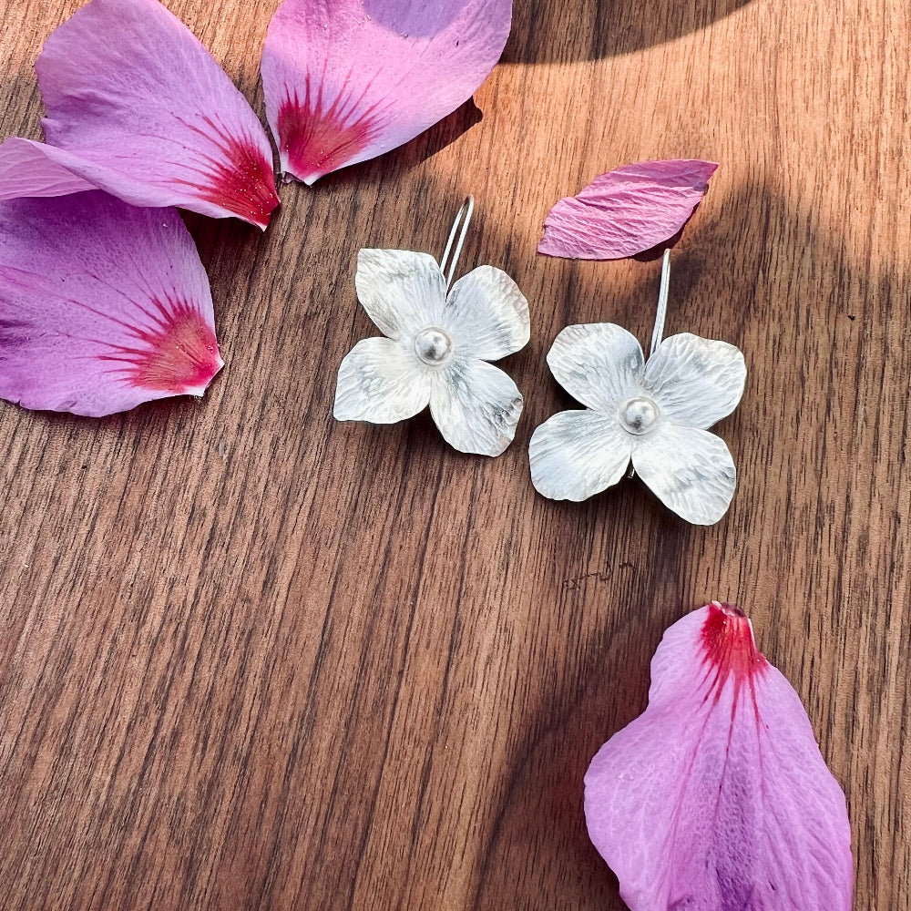 Flower-shaped silver earrings on a wooden surface with pink petals.
