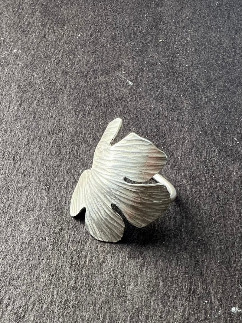 Silver fig leaf-shaped ring on a dark gray surface. Wraps around finger and has hammered detailing.