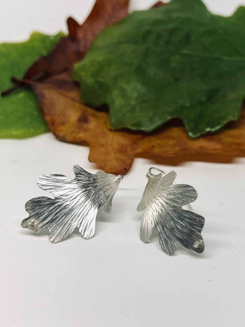 Silver leaf-shaped earrings on a white surface with green leaves and brown leaves in the background.