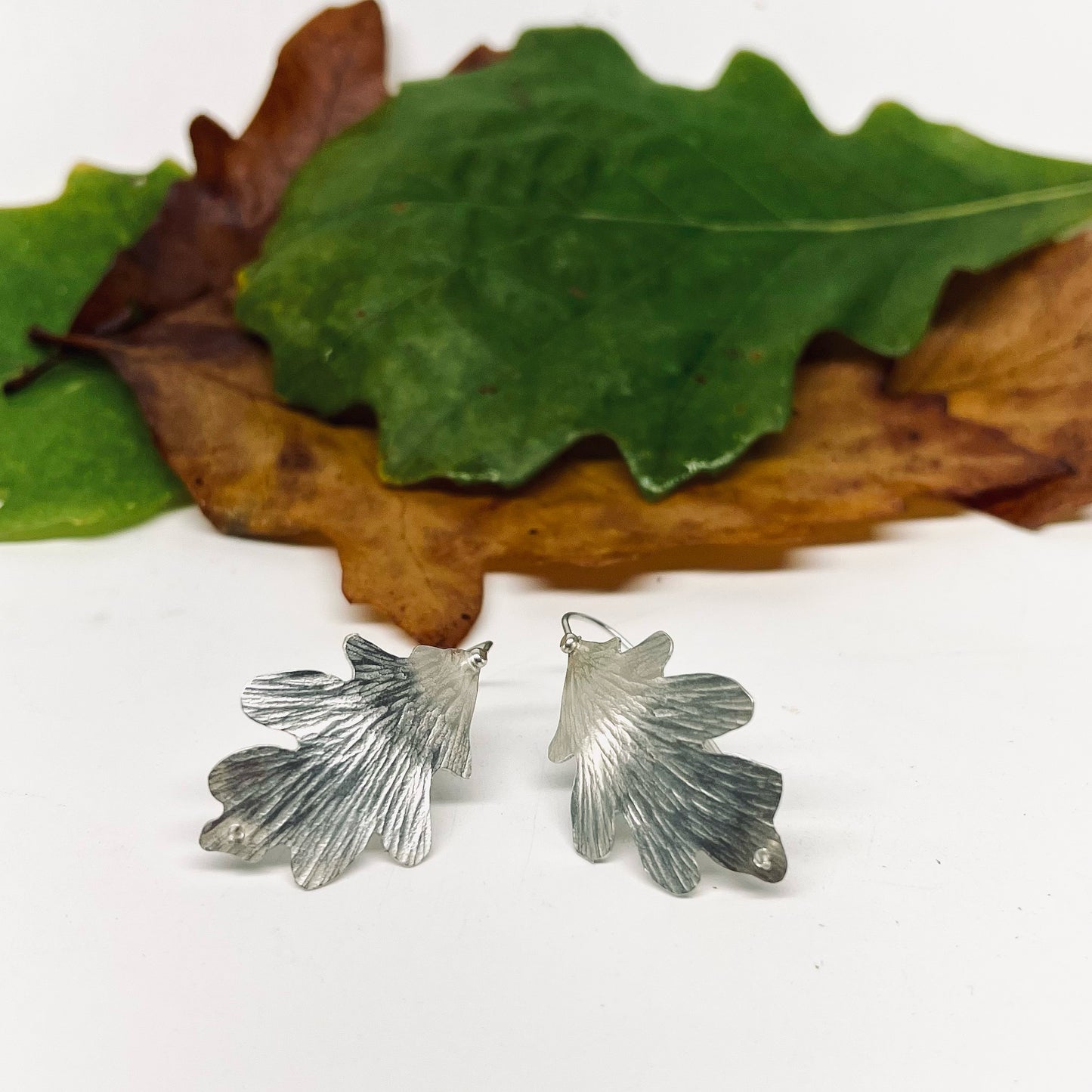 Silver leaf-shaped earrings on a white surface with green and brown leaves.