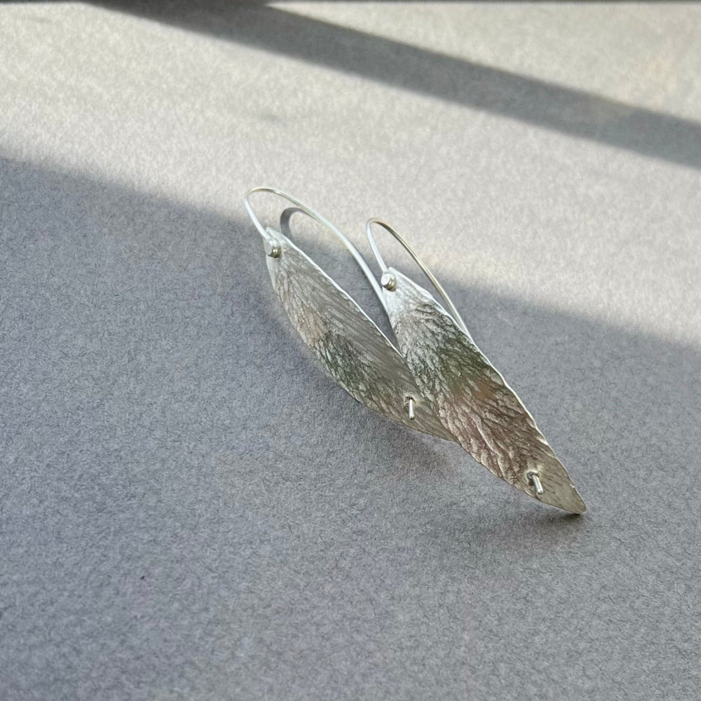 Silver leaf-shaped earrings on a light gray surface with soft shadows